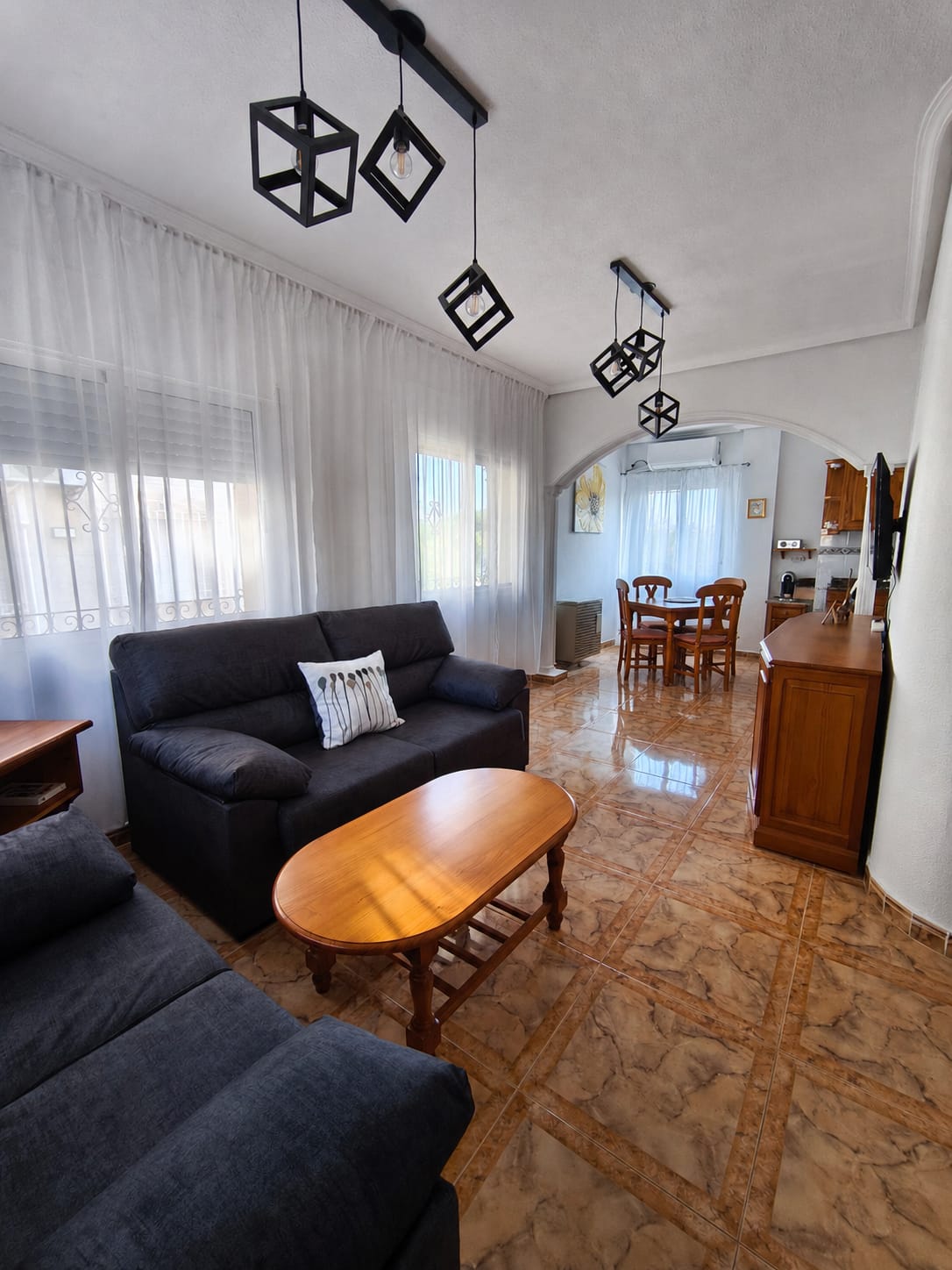 Open-plan living/dining area with two dark sofas, coffee table, geometric pendant lights, archway leading to dining area with wooden table and chairs, sheer curtains, and marble-effect floor tiles
