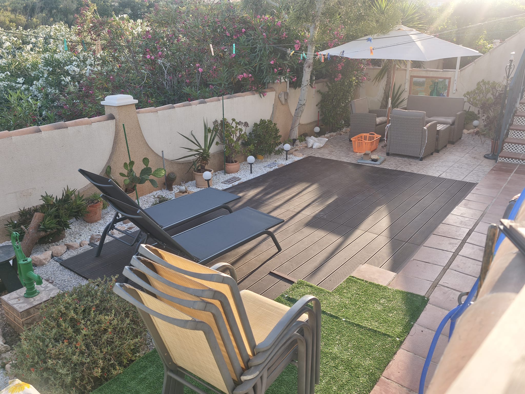 Sunny garden with sun loungers, composite decking, rattan seating under parasol, cacti, globe lanterns, and flowering oleander